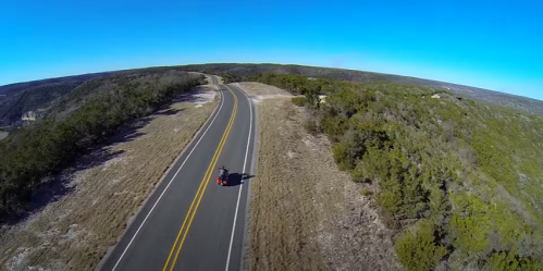 Aerial view of a winding road surrounded by green hills and clear blue sky. A motorcycle is seen on the road.