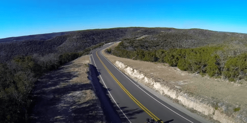 A winding road cuts through a hilly landscape under a clear blue sky, surrounded by greenery and rocky terrain.