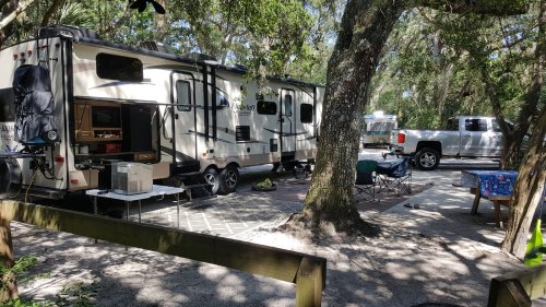 A camping scene featuring a trailer, a pickup truck, and outdoor seating under trees in a shaded area.