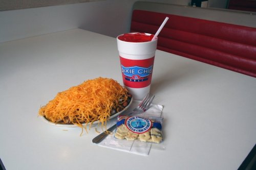A plate of cheese-covered chili with a drink and a packet of crackers on a table.