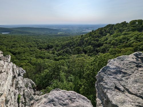 A panoramic view of lush green hills and valleys under a clear sky, framed by rocky outcrops in the foreground.