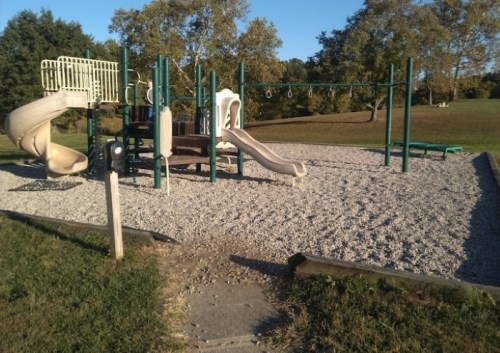 Playground with slides, swings, and climbing structures on a gravel surface, surrounded by grass and trees.