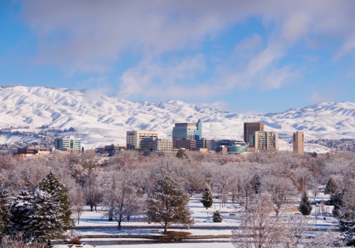 Snow-covered landscape with a city skyline in the background, surrounded by mountains under a clear blue sky.