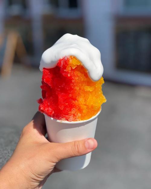 A hand holding a cup of colorful shaved ice topped with white cream, featuring red and orange layers.
