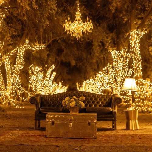 A cozy outdoor scene with a black couch, a vintage trunk, a lamp, and a chandelier, all surrounded by warm fairy lights.