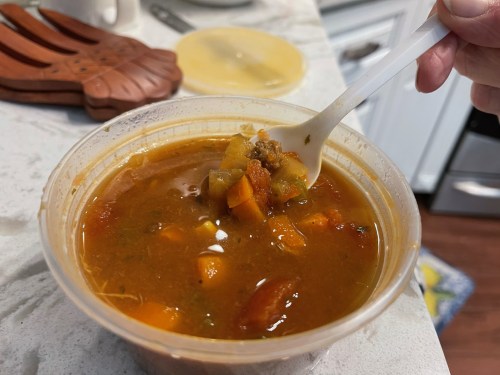 A hand holding a spoon with a bite of vegetable soup from a plastic container on a kitchen countertop.