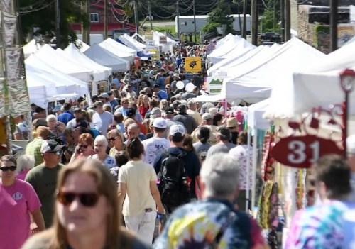 A bustling outdoor market with numerous tents and a large crowd of people walking through the street.