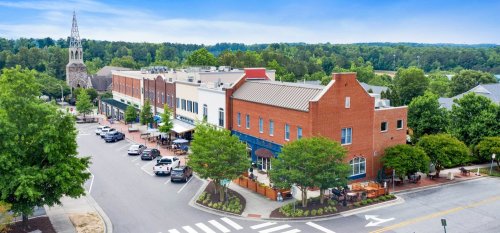 Aerial view of a charming town square with shops, trees, and a church steeple in the background.