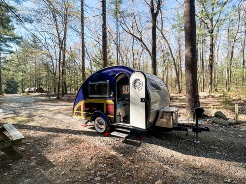 A colorful teardrop camper parked in a wooded area, surrounded by trees and a gravel path.