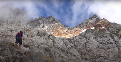 A hiker ascends rocky terrain under a dramatic sky with mountains in the background.