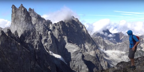 A person in a blue helmet and colorful shirt gazes at rugged mountain peaks under a clear sky with scattered clouds.