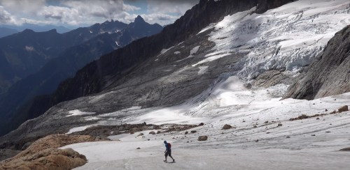 A lone hiker traverses a snowy glacier surrounded by rugged mountains under a partly cloudy sky.