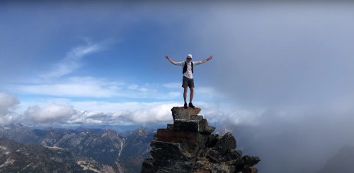A person stands triumphantly on a rocky peak, arms outstretched, with mountains and clouds in the background.