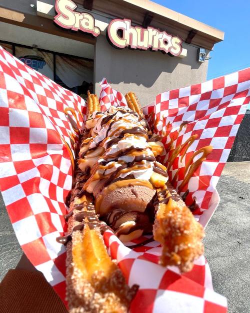 A large churro topped with ice cream, whipped cream, chocolate, and caramel sauce, served in a red checkered paper.