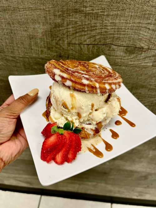 A hand holding a plate with a dessert featuring ice cream between two cinnamon rolls, drizzled with caramel and garnished with strawberries.
