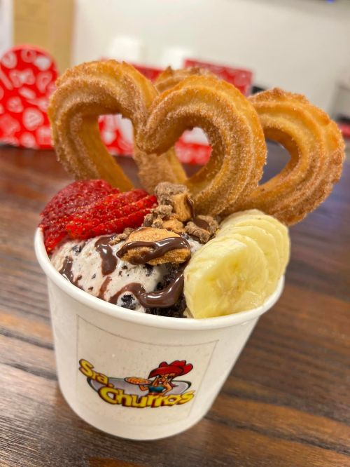 A cup of ice cream topped with banana slices, strawberries, chocolate drizzle, nuts, and heart-shaped churros.
