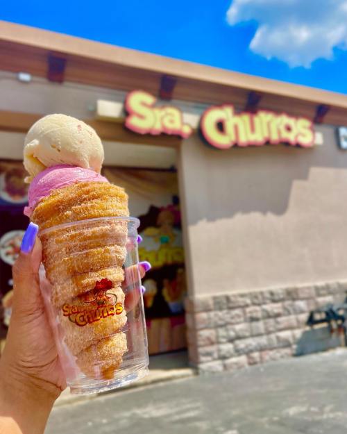 A hand holds a cone made of churros filled with ice cream, in front of a shop sign that reads "Sra. Churros."