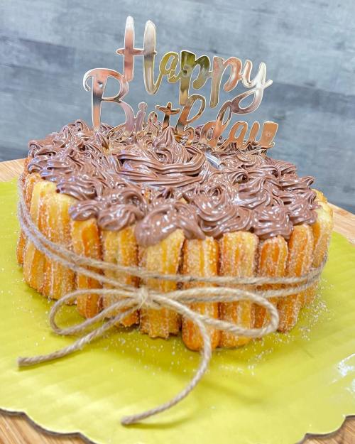 A round birthday cake decorated with chocolate frosting, churros, and a "Happy Birthday" topper, tied with twine.