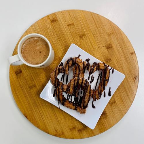 A plate of chocolate-drizzled churros next to a cup of coffee on a wooden surface.