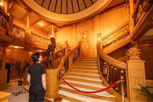 A person gazes up at an ornate staircase in a grand, wooden interior with decorative railings and a clock.