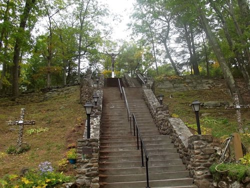 Stone steps lead up a hillside, flanked by trees and lamps, with a cross at the top surrounded by greenery.
