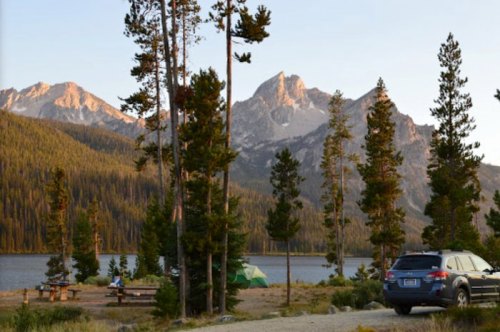 Scenic view of mountains and a lake, with trees, a campsite, and a parked car in the foreground during sunset.