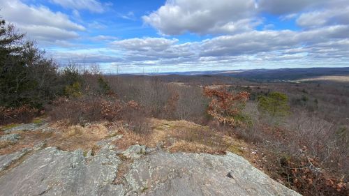 A rocky outcrop overlooks a vast landscape of rolling hills and a cloudy blue sky.
