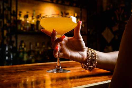 A hand holding a yellow cocktail in a coupe glass, set against a dimly lit bar with bottles in the background.