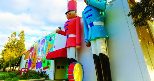 Two large toy soldier figures stand outside a colorful mural on a building, surrounded by greenery.