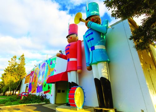 Two large toy soldier sculptures stand outside a colorful mural on a building, with a blue sky and trees in the background.
