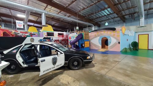 A colorful indoor play area featuring a police car, slides, and whimsical murals on the walls.