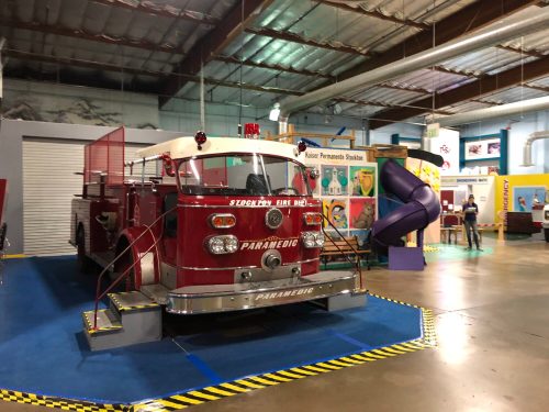 A vintage fire truck displayed in a colorful indoor exhibit space, with a slide and various educational displays in the background.