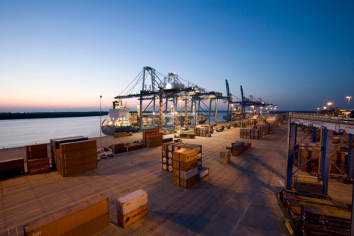 Container ships and cranes at a port during twilight, with stacked cargo containers on the dock.