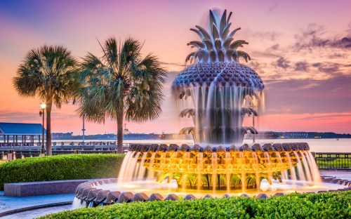 A decorative pineapple fountain surrounded by palm trees at sunset, with water cascading and a colorful sky in the background.