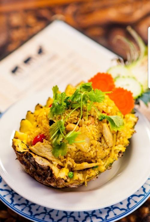 A vibrant dish of fried rice served in a pineapple, garnished with cilantro, carrots, and cucumber slices.
