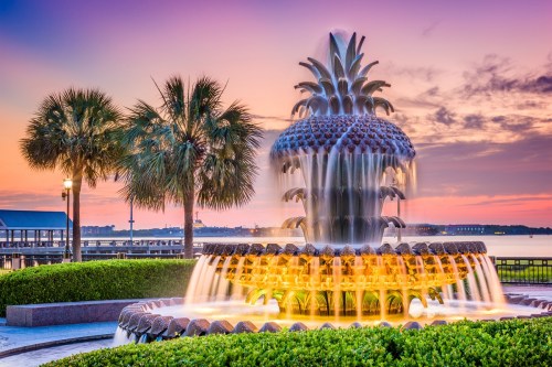 A pineapple-shaped fountain surrounded by palm trees at sunset, with water cascading and a colorful sky in the background.