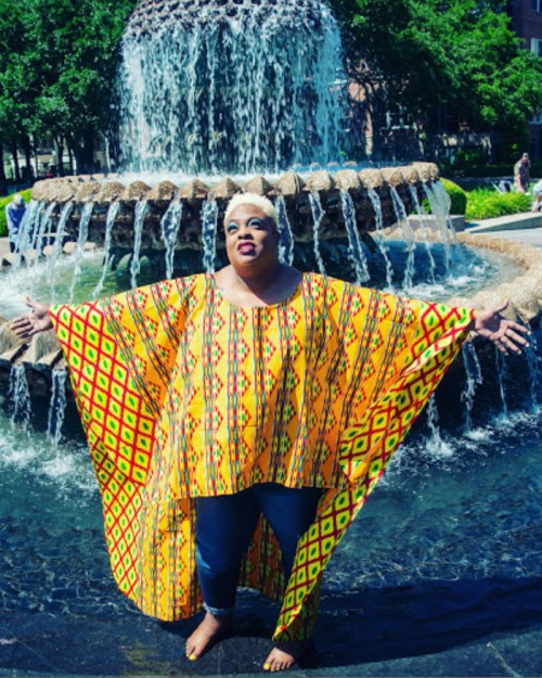 A person in a vibrant yellow patterned outfit stands in a fountain, arms outstretched, with water cascading around them.