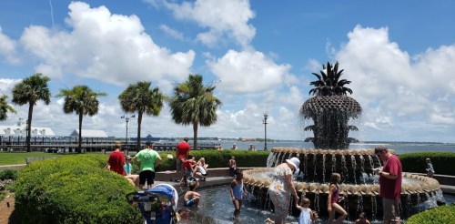 Children and adults play around a large fountain on a sunny day, with palm trees and a waterfront in the background.
