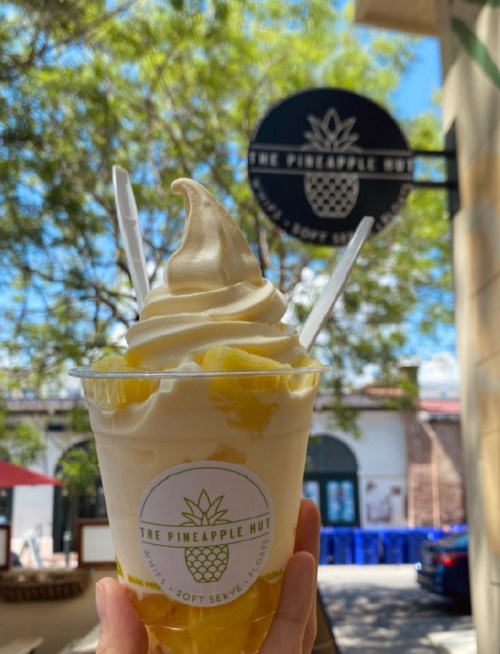 A hand holds a cup of soft serve ice cream topped with pineapple chunks, with "The Pineapple Hut" sign in the background.