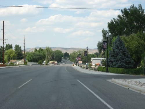A wide, empty road lined with trees and lampposts, leading towards distant hills under a partly cloudy sky.