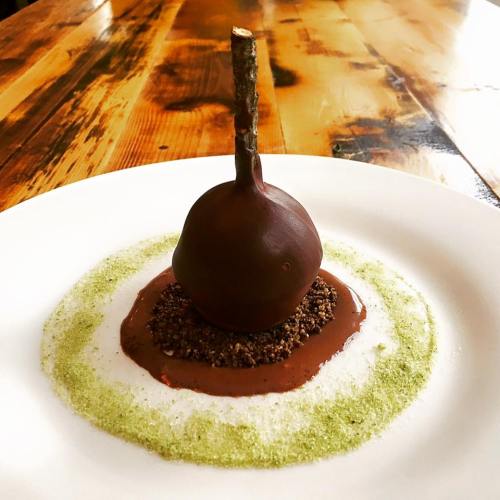 A chocolate dessert shaped like a pear, resting on a circular sauce with green and brown elements on a white plate.