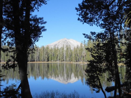A serene lake reflects a mountain, framed by tall trees under a clear blue sky.