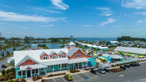 Aerial view of a colorful waterfront resort with pools, green spaces, and parked vehicles under a clear blue sky.