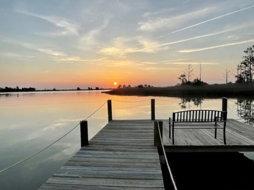 A serene sunset over calm waters, viewed from a wooden dock with a bench, surrounded by trees and soft clouds.