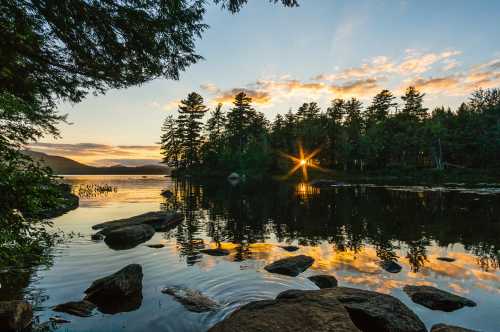 Sunset over a tranquil lake, with trees reflecting on the water and rocks in the foreground.