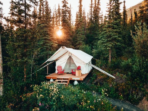 A cozy glamping tent with red chairs, surrounded by trees and wildflowers, illuminated by a sunset.