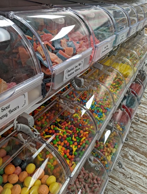 A colorful display of various candies in clear bins, labeled with names, arranged neatly on a wooden shelf.
