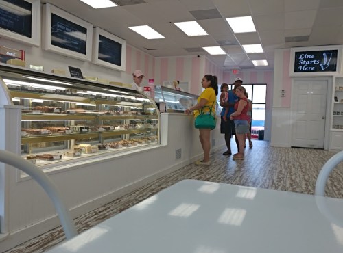 A bright bakery interior with a display case of pastries and a line of customers waiting to order.