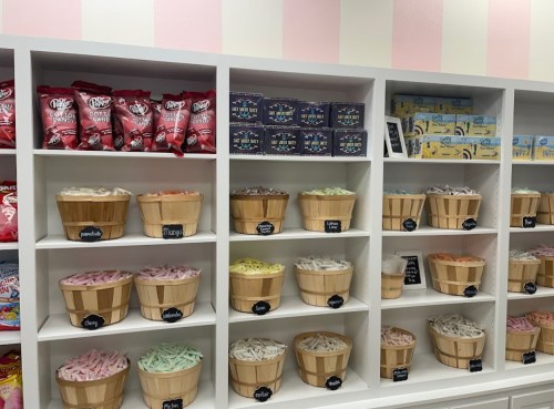 Colorful candy display with wooden baskets filled with various sweets on white shelves against a pink-striped wall.