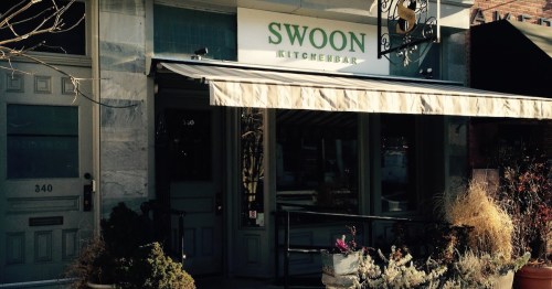 Exterior of Swoon Kitchenbar, featuring a sign and awning, with plants and a welcoming entrance.